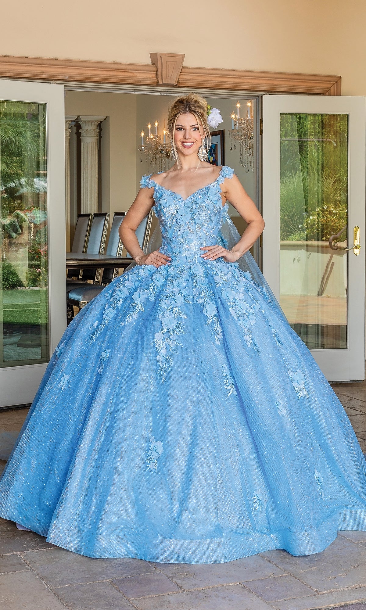 A woman stands outside double doors wearing the Quinceanera Dress 1707 by Dancing Queen, a voluminous light blue off-the-shoulder ball gown adorned with floral embroidered appliqués.